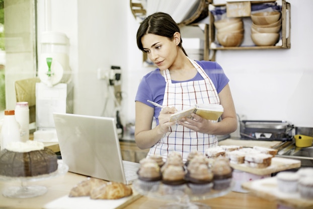Woman writes in a notebook while looking at her laptop. She is in a kitchen surrounded by home-baked goods.