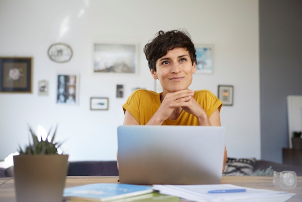 A woman is seated at her kitchen table. She has her laptop in front of her plus some books and papers spread out before her. She has a slight smile on her face. She is planning her home-based business.