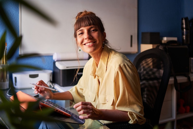 A woman graphic artist poses in her home studio at the viewer.