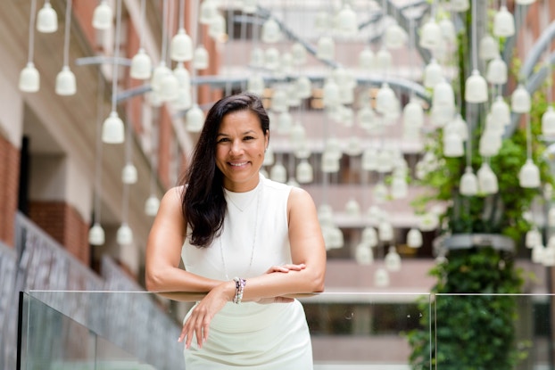 An image of Tiffany Houser, founder and CEO of EvolveEQ, leaning on a glass panel in the outdoor atrium of a multi-story building. Tiffany is a smiling woman with long, dark hair draped over one shoulder. She wears an ivory sleeveless top and a long, thin chain necklace.