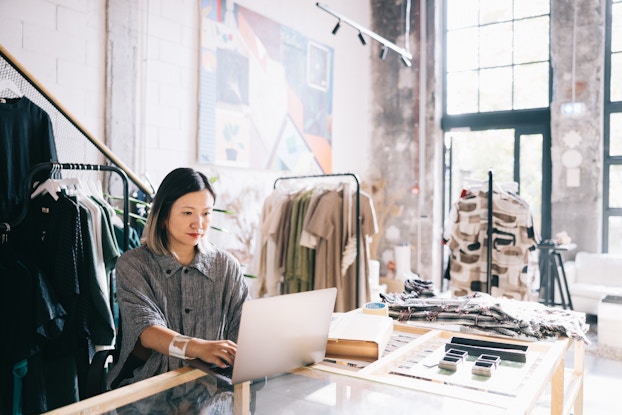 A woman in a large, high-ceilinged workroom sits at a worktable and types on a laptop. The woman has shoulder-length dark hair with light streaks, and she wears a loose-fitting gray button-up shirt and a silver cuff bracelet. Around the room are racks of neutral-colored clothing, and swatches of fabric are piled on the worktable.