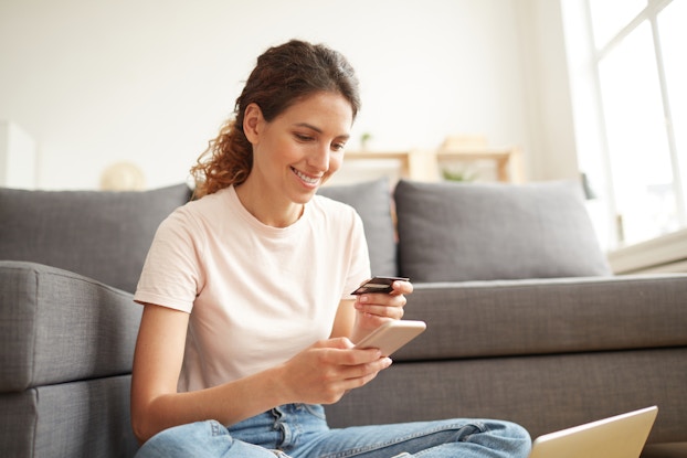 A young woman with her hair in a ponytail sits on the floor in front of a gray couch. She holds a smartphone in one hand and a credit card in the other, and looks down at both with a smile.