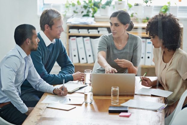 Four people sit around a small conference table in an office. The woman sitting at the head of the table is speaking and gesturing to an open laptop. The other three people around the table, two men and a woman, are watching her with pens in their hands. In the background are shelves filled with thick binders.