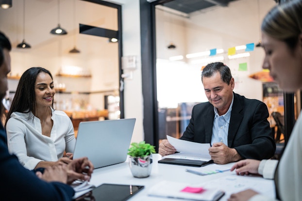 A group of businesspeople are seated around a conference room table. One of the individuals, a man, reviews accounting records.