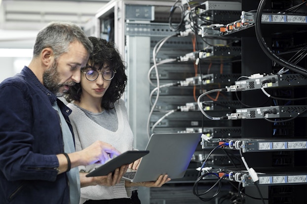 Two people stand next to a bank of computer servers. The person on the left, who is holding a small electronic tablet, is an older man with graying hair and a salt-and-pepper beard. He is pointing to something on the tablet's screen. The person on the right is a younger woman, with shoulder-length dark curly hair and glasses; she holds an open laptop.