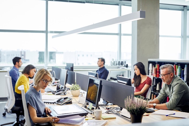 A brightly lit, modern office is shown. There are six work professionals seated at a long table in an open-office layout working on their desktop computers.