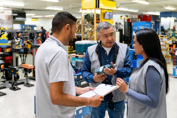 The owner of a hardware store trains two new employees while holding a drill.