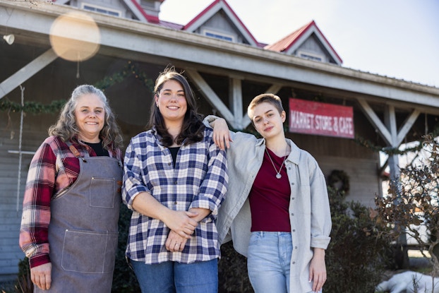 A woman business owner of a market and cafe stands in front of her business with her daughter and grandchild.