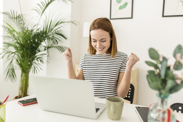 Woman working from home on her laptop, excited with her hands in the air.