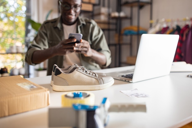 A pair of pale gray sneakers sits on a table next to an open laptop, a shipping box, and a few office supplies. In the background, slightly out of focus, a young man takes a picture of the shoes with his smart phone.
