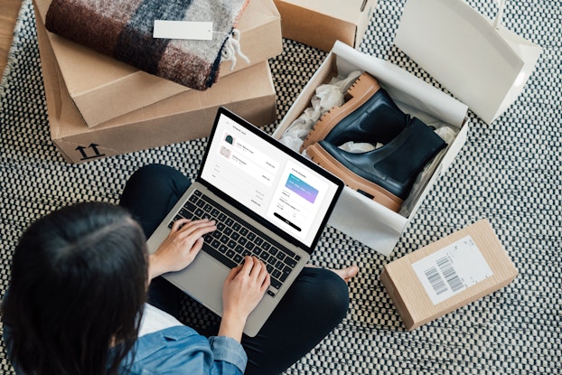 Woman sitting on the floor next to shoeboxes, shopping on her laptop.