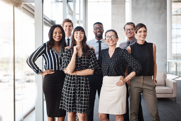 A diverse group of people in businesswear stand in a large open office space next to floor-to-ceiling windows.