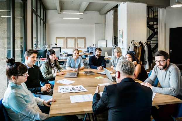A group of individuals are pictured around a large conference table for a meeting. The setting is a modern office.