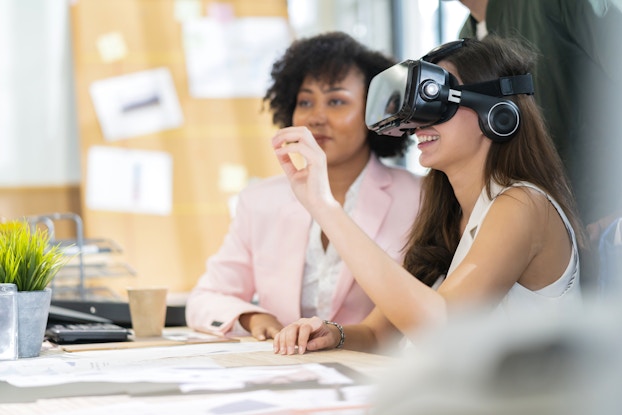 A woman wearing a VR headset sits at a desk and extends one hand in front of her, reaching to touch something that only she can see. Another woman sits beside her and someone else stands behind her; the latter person's head is cut off by the edge of the picture. The room around them appears to be an office or a staff room at a school; a large bulletin board can be seen out of focus in the background.
