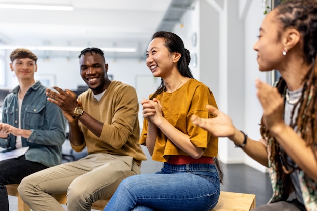 Four people sit on wooden benches in a large open room. The four people (a person in a denim jacket, a man in a tan sweater and khakis, a woman in a goldenrod blouse and jeans, and a woman with long hair who sits on the far right, out-of-focus and partially outside of the image) are applauding and smiling. The group's focus is on the woman in the goldenrod shirt, who has her hands clasped together in joy.