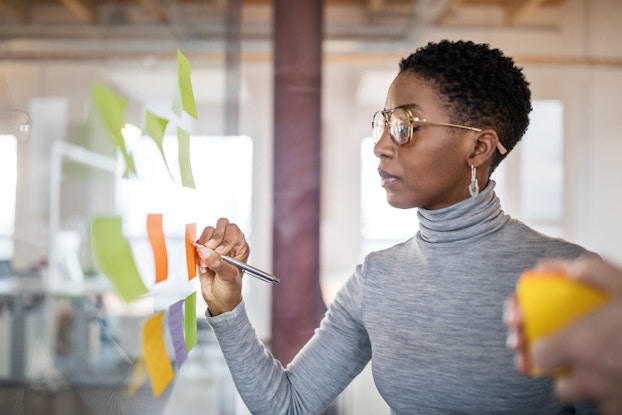 A businesswoman writes new ideas onto a adhesive note affixed to a glass wall.