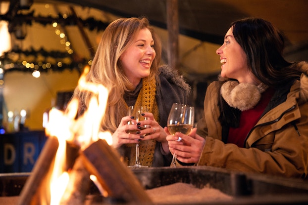 Two women friends sit before a fire pit. They are laughing and are each holding a glass of wine in their hands.