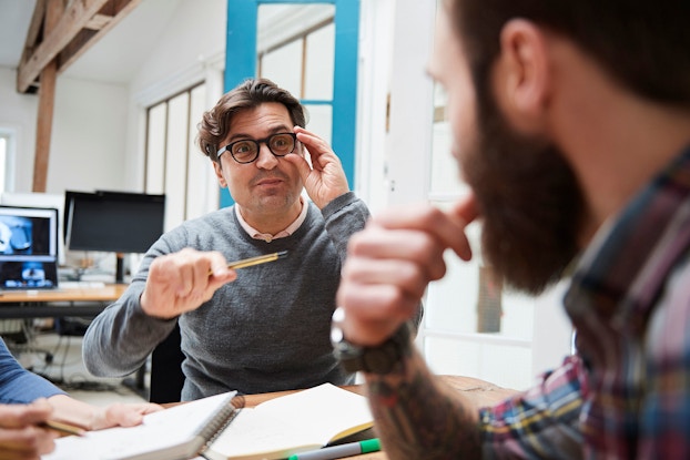 A business owner and a male employee sit in an office brainstorming how to solve a problem. The male business owner is gesturing with his hand to make a point.