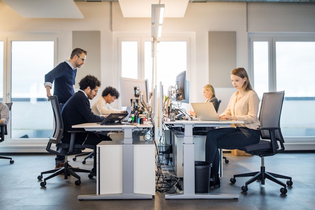 Five people are seated at desks in an open-office environment working on their laptops while a manager is standing and talking to one employee.