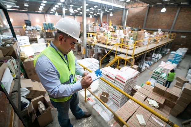 A safety inspector in a white hard hat stands on a plaform overlooking at large warehouse and writes something on a paper attached to a clipboard. The inspector is a man in a light blue button-down shirt, blue jeans, and a neon green high-visibility vest. He stands behind a yellow railing and in front of shelves overflowing with cardboard boxes.