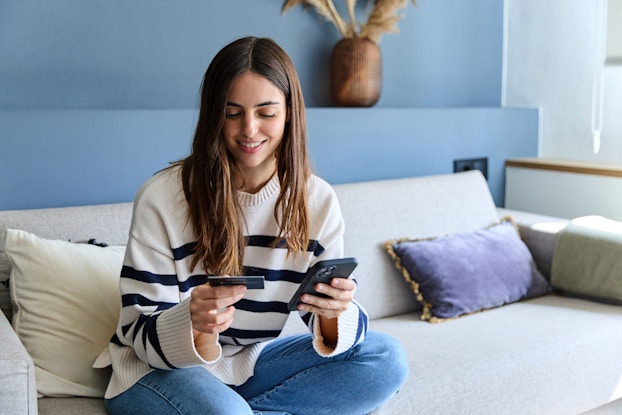 A young woman is seated on a couch in her house. She is holding a credit card in her right hand and her mobile phone in her left hand. She is making an online purchase.