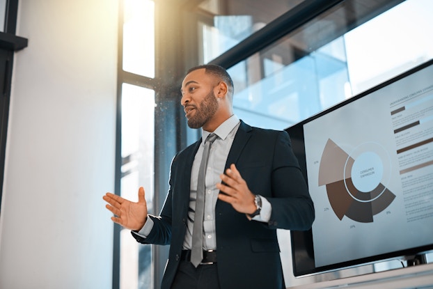 A man in a suit stands in front of a floor-to-ceiling window and speaks and gesturing to a group off-camera. Behind him is a screen showing a graph in various shades of brown.