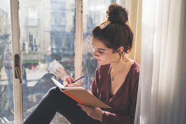 A young woman sits in a window seat and writes in a journal. The woman has dark hair pulled into a messy bun, and she wears dark jeans, a loose-fitting maroon blouse, and a silver locket.