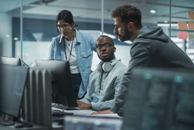 Three people with serious expressions sit and stand around a pair of computer monitors, which are turned away from the viewer. The man sitting down has a pair of white headphones hanging around his neck. He looks up at another man, who wears a hoodie and leans over the desk. On the other side of the seated man is a woman in a chambray shirt and glasses; she looks at one of the monitors. In the foreground are more computer monitors, out of focus.