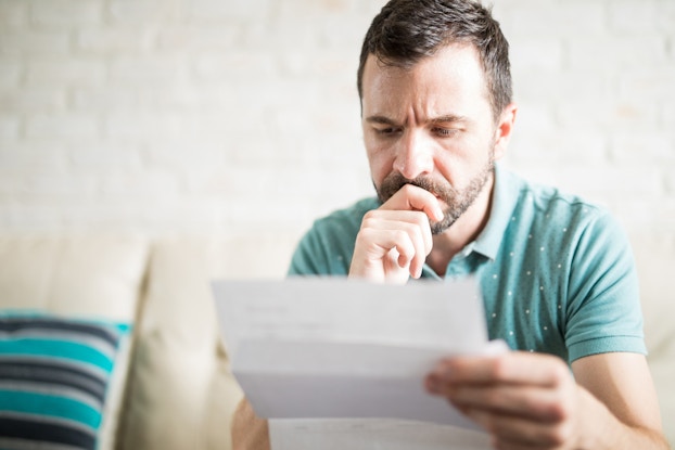 A bearded man sits on a cream-colored sofa and looks at a creased piece of paper with a worried expression.