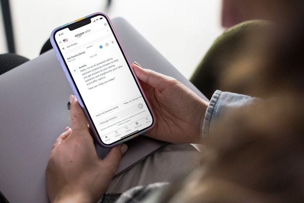A pair of hands hold a smartphone, the screen of which shows the page for Amelia, Amazon's generative AI-based sales assistant. The Amelia chatbot has typed "Hello, I'm an AI-powered selling assistant created by Amazon to help you get answers to your selling questions and insights into your sales and traffic metrics. How can I help you today?"