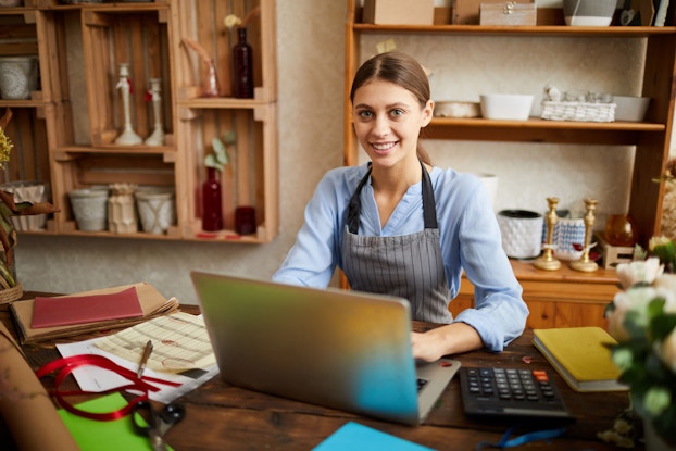 A woman small business owner is pictured standing at a table in her store. She is using laptop and looking at the camera.