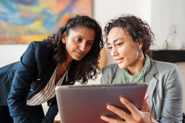 Two women business professionals look at a computer tablet. They are searching a computer database to locate possibilities for the name of their business.
