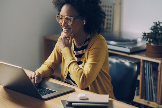 Woman in glasses smiles as she works on a laptop in her home.