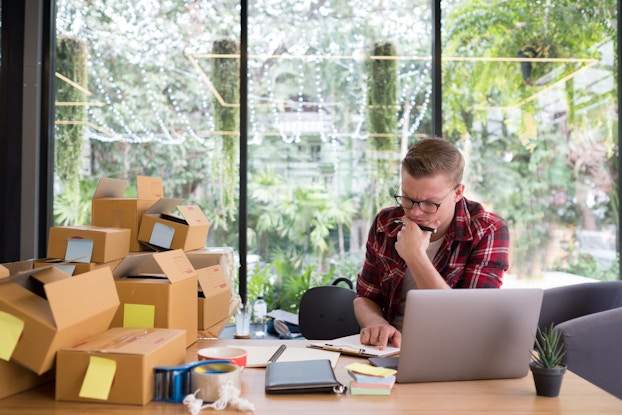 Entrepreneur multitasks in a cluttered home office, boxes on desk next to his workspace