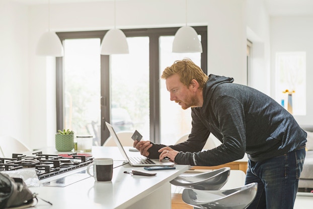 A man stands in his kitchen with a laptop sitting on the counter. The man makes an online purchase using a credit card, which he is holding in his right hand.