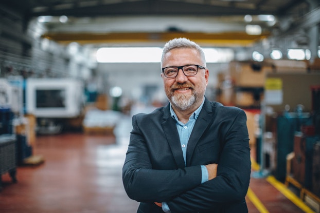 The CEO or general manager of a manufacturing company is pictured smiling at the viewer with his arms folded. Behind him in the background is the factory he manages.