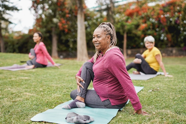 An older woman sits outside, on a light teal yoga mat stretched out over the grass. She has her right leg crossed over her left leg in the "Lord of the Fishes" pose. The woman is smiling and wearing gray yoga pants, a pink long-sleeved shirt, and gray-and-white ankle socks. Behind her, out of focus, are two more women: a short-haired woman in a yellow shirt and a dark-haired woman in a pink shirt. A line of trees stands at the edge of the grass.