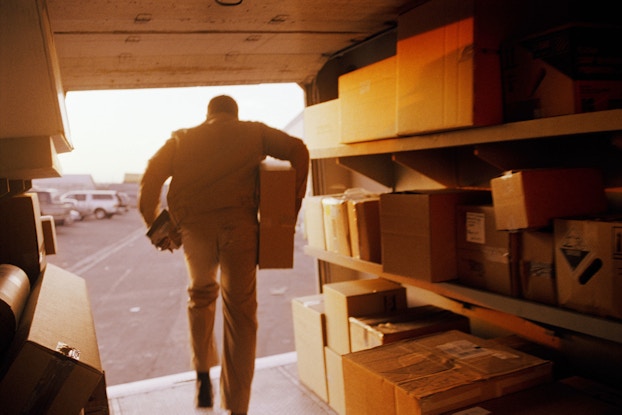 A freight delivery driver exits the back of his truck. He has a box under his right arm and he is carrying a bar code scanner in his left hand.
