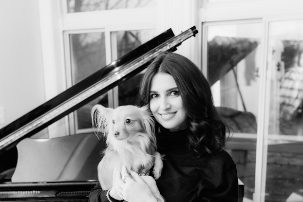 A black-and-white photo of Shahla Karimi, Founder of Shahla Karimi Jewelry, standing in front of a baby grand piano and holdling a small dog. Shahla is a young woman with long dark hair wearing a dark-colored sweater and smiling for the camera.