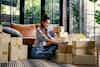 A woman sits cross-legged on a living room floor surrounded by taped-up cardboard boxes. She types something on a laptop perched on top of two small boxes. Behind her is a brown leather couch; behind the couch is a set of floor-to-ceiling windows, through which can be seen a yard filled with tall leafy ferns.
