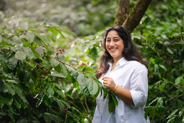 A photo of Shonali Paul, Founder of Paul John Indian Caffeine Company, standing outside surrounded by leafy bushes. Shonali has long dark hair and wears a white button-up shirt, a gold bracelet, and a gold pendant necklace. She has sunglasses perched on top of her head and a smile on her face.