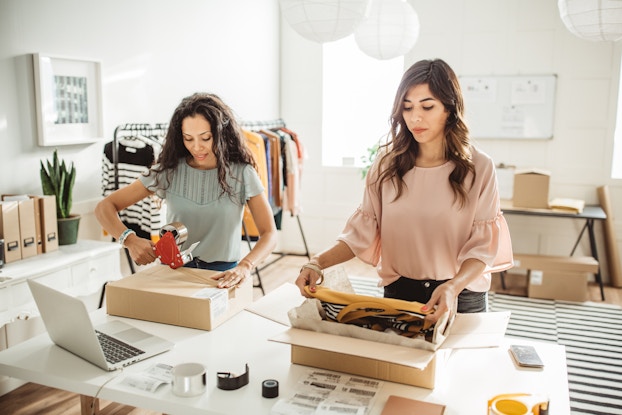 Two young women pack boxes with merchandise. Across from them on the table is an open laptop.