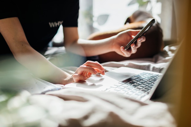 A close-up of a person seen from the neck-down using a laptop. The person uses one hand to control the laptop's trackpad, while their other hand holds a smartphone. The person wears a dark T-shirt with the word "WHY" written on the right side of the chest.