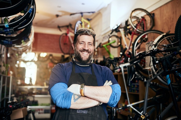 A bearded man stands in a storeroom, the walls of which are covered with handing and mounted bikes. The man wears a black apron over layered blue shirts and he has a tattoo of a triangle on one arm. The man has his arms crossed and smiles proudly.