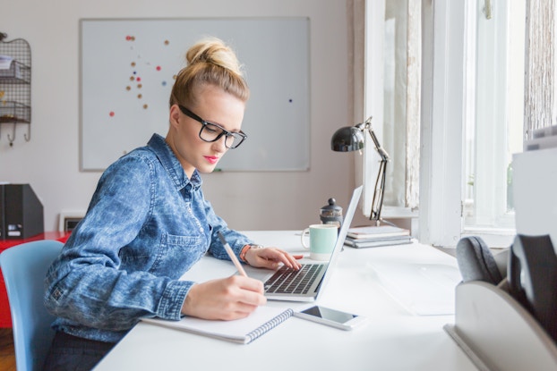 A young blonde woman sits at a desk with one hand on the keyboard of an open laptop and the other hand using a pen to write on a pad of paper. The woman is wearing a denim shirt and glasses, and she has her hair piled on top of her head in a messy bun. In the background is a white office room with a large whiteboard on one wall. The woman's desk is next to a large set of windows, so the light from outside falls across the woman as she works.