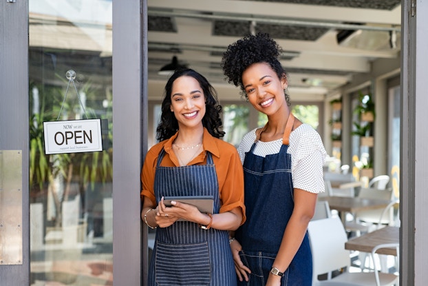Two women stand smiling in the doorway of a restaurant or cafe. The space behind them is filled with square wooden tables and white chairs; baskets with plants growing out of them are mounted on the walls. The woman on the left wears an orange collared shirt under a dark-blue-and-white-striped apron. She holds a digital tablet. To the left of her is the open door, which has an open sign hanging from a suction cup on its window. The woman on the right is slightly taller and wears a white T-shirt under a dark blue apron.