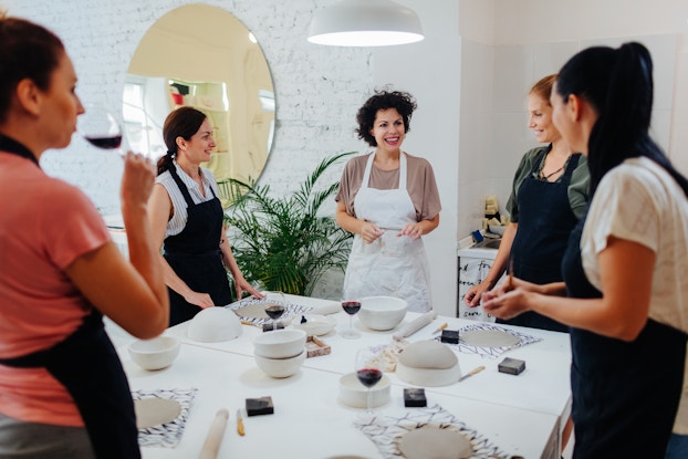 A group of women in aprons stand around a table in a large white room. Each woman has a circle of gray clay sitting on a placemat in front of her on the table; the table also holds several white ceramic bowls, a few carving tools, and enough glasses of red wine for everyone. The woman closest to the camera, standing in the foreground and facing away from the viewer, is taking a sip of wine. Most of the women are wearing black aprons, but the woman in the center of the image is wearing a white one. The other women are turned to face the woman in the white apron, who is smiling at the group.
