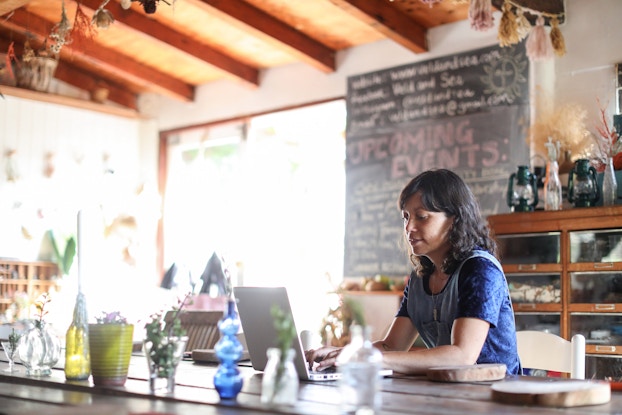 A wide shot of a woman sitting at a wide table in a public community space, typing on a laptop. The woman has shoulder length dark hair and is wearing a dark blue T-shirt under a light blue tank top. The room she's in is warmly lit with exposed beams on the ceiling and a blackboard taking up part of one wall. The blackboard has "UPCOMING EVENTS" written in large letters with red chalk, with smaller writing in yellow above and below. On the table across from the woman is a row of glass vases and jars in various colors and shapes.