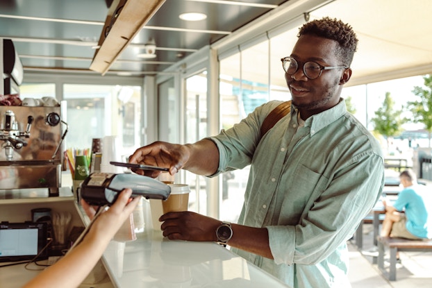 A man in glasses and a button-up shirt uses his smartphone to scan a handheld payment device at a cafe. The person holding the POS device is behind the cafe's service counter; only their arm can be seen.