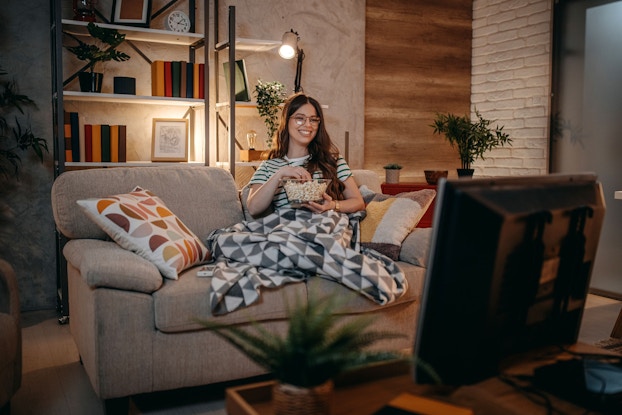 A young woman is watching TV at night. She is sitting on a couch in her home's living room. She is eating popcorn and is wrapped in a blanket.